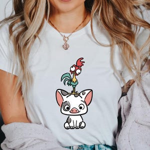 Puede incluir: Camiseta blanca con una ilustraci&oacute;n de dibujos animados de un cerdo y un gallo. El cerdo es blanco con manchas negras y orejas rosas. El gallo es colorido con acentos rojos, verdes y amarillos. Prenda de vestir informal.