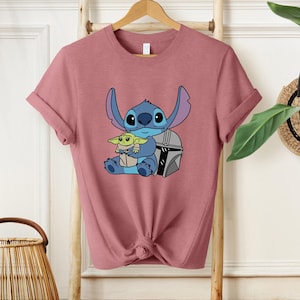May include: A dusty rose t-shirt featuring a graphic of Stitch and Grogu from Star Wars. Stitch is blue with large ears, and Grogu is green. The design includes a silver helmet. The shirt has rolled sleeves and a knotted hem.