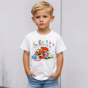May include: A white t-shirt featuring a colorful cartoon design of various cars, a birthday cake, and balloons. The shirt has the number "2" and the name "Name" printed on it. The shirt is being worn by a young child.