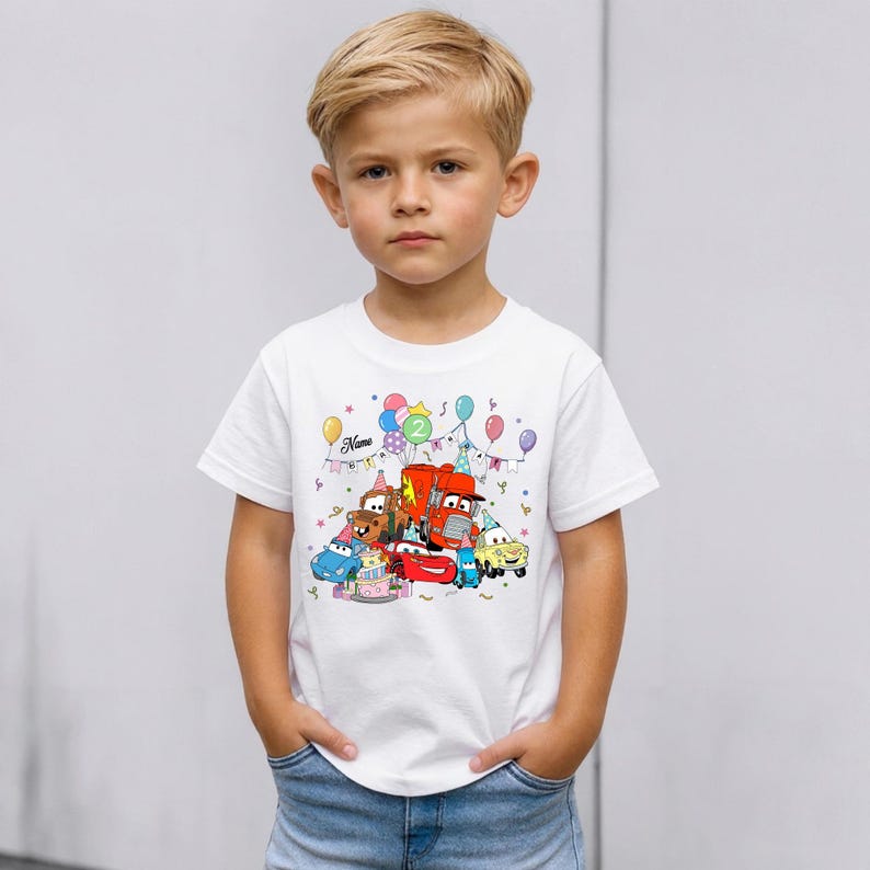 May include: A white t-shirt featuring a colorful cartoon design of various cars, a birthday cake, and balloons. The shirt has the number "2" and the name "Name" printed on it. The shirt is being worn by a young child.