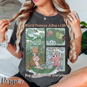 May include: Dark grey t-shirt featuring a colorful graphic from "A Bug's Life." The design includes cartoon images of insects in various scenes, with the text "World Famous A Bug's Life" and "Bug-sized Fun For Everyone."