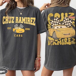 May include: Dark grey t-shirt featuring a yellow graphic of Cruz Ramirez from the Cars movie. The front design includes the text "CRUZ RAMIREZ" and "CARS", with a yellow car and checkered flags. The back has a larger design of the car.