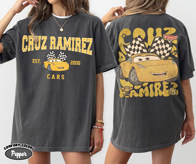 May include: Dark grey t-shirt featuring a yellow graphic of Cruz Ramirez from the Cars movie. The front design includes the text "CRUZ RAMIREZ" and "CARS", with a yellow car and checkered flags. The back has a larger design of the car.