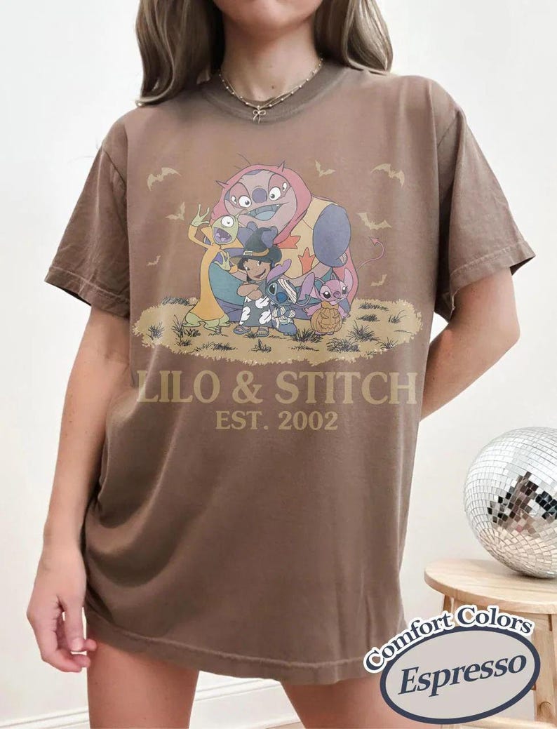 Op de afbeelding: Espresso-kleurige t-shirt met een afbeelding van Lilo & Stitch-personages, met de tekst "LILO & STITCH EST. 2002." Het ontwerp bevat stripfiguren en vleermuizen. Het shirt is van Comfort Colors.