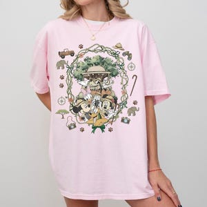 Puede incluir: Camiseta rosa claro con un gr&aacute;fico colorido de personajes de Disney con atuendo de safari. El dise&ntilde;o incluye a Mickey Mouse, Minnie Mouse, Donald y Goofy, rodeados de ilustraciones tem&aacute;ticas de safari como un jeep, elefantes y huellas de patas.
