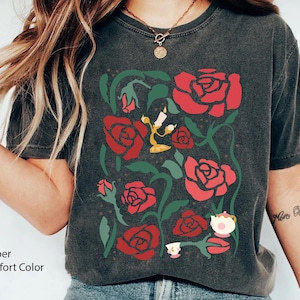 Puede incluir: Camiseta gris oscuro con un diseño floral con rosas rojas, enredaderas verdes e ilustraciones de dibujos animados de un candelabro y una taza de té. La camiseta tiene las palabras "Pepper" y "Comfort Color".