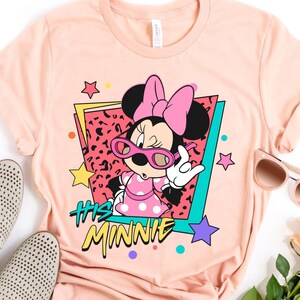 K&ouml;nnte beinhalten: Ein pfirsichfarbenes T-Shirt mit einer Grafik von Minnie Mouse mit rosa Sonnenbrille und einem gepunkteten Kleid. Das Design enth&auml;lt den Text "His Minnie" in einem Retro-Stil mit Sternen und farbigen Akzenten.