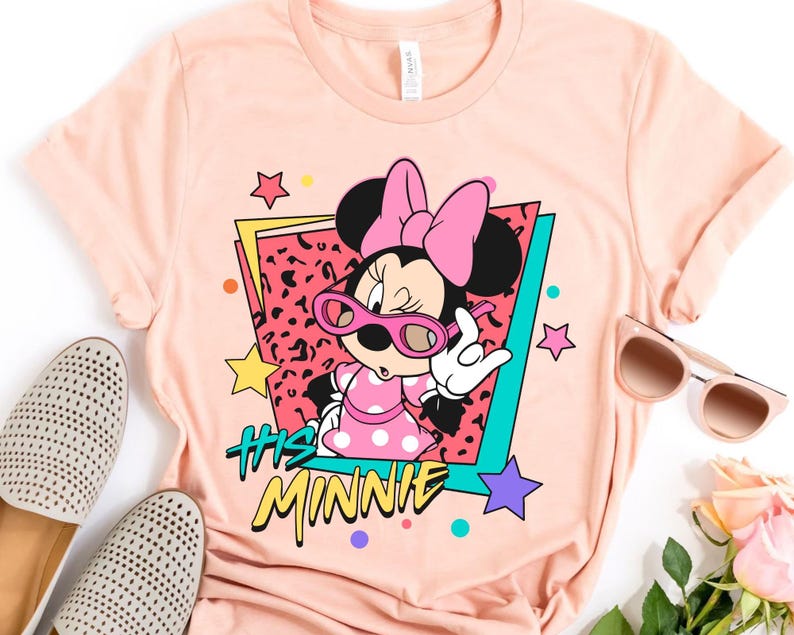 K&ouml;nnte beinhalten: Ein pfirsichfarbenes T-Shirt mit einer Grafik von Minnie Mouse mit rosa Sonnenbrille und einem gepunkteten Kleid. Das Design enth&auml;lt den Text "His Minnie" in einem Retro-Stil mit Sternen und farbigen Akzenten.