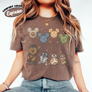 May include: Espresso-colored t-shirt featuring Star Wars characters and Mickey Mouse-shaped balloons. Chewbacca, R2-D2, BB-8, and an Ewok are depicted below the balloons. The shirt has the words "Comfort Colors" and "Espresso" in a circular design.