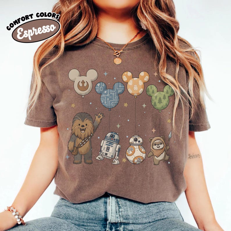 May include: Espresso-colored t-shirt featuring Star Wars characters and Mickey Mouse-shaped balloons. Chewbacca, R2-D2, BB-8, and an Ewok are depicted below the balloons. The shirt has the words "Comfort Colors" and "Espresso" in a circular design.
