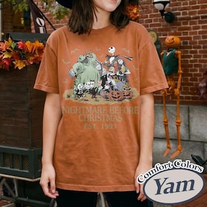 Puede incluir: Una camiseta de color naranja quemado con un gr&aacute;fico de "Pesadilla antes de Navidad". El dise&ntilde;o incluye personajes como Jack Skellington, Sally y Oogie Boogie, con el texto "NIGHTMARE BEFORE CHRISTMAS EST. 1993". La camiseta tiene una etiqueta Comfort Colors.