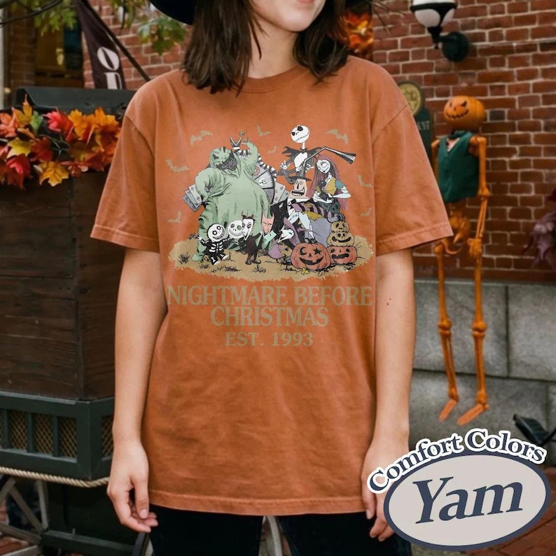 Puede incluir: Una camiseta de color naranja quemado con un gr&aacute;fico de "Pesadilla antes de Navidad". El dise&ntilde;o incluye personajes como Jack Skellington, Sally y Oogie Boogie, con el texto "NIGHTMARE BEFORE CHRISTMAS EST. 1993". La camiseta tiene una etiqueta Comfort Colors.