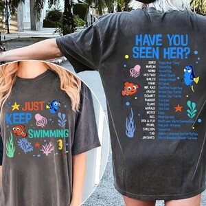 May include: Dark gray t-shirt featuring the phrase "Just Keep Swimming" with cartoon sea creatures. The back of the shirt has the text "Have You Seen Her?" and a list of characters from a popular animated film.