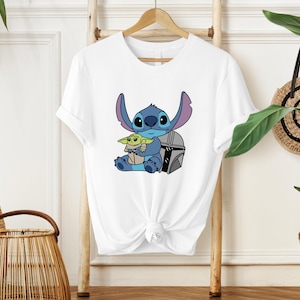 May include: White t-shirt featuring a cartoon illustration of Stitch and Grogu. Stitch is blue with large ears, and Grogu is green. They are sitting together with a silver helmet. The t-shirt is hanging on a wooden hanger.