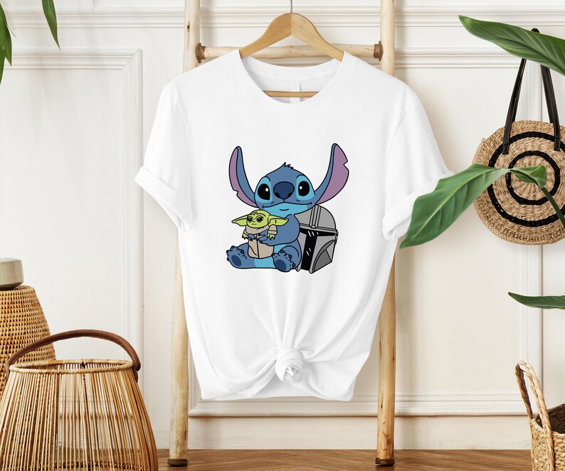 May include: White t-shirt featuring a cartoon illustration of Stitch and Grogu. Stitch is blue with large ears, and Grogu is green. They are sitting together with a silver helmet. The t-shirt is hanging on a wooden hanger.