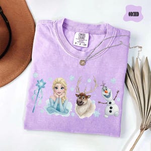 Peut inclure: T-shirt lilas avec Elsa, Olaf et Sven de La Reine des Neiges. Le t-shirt a un col rond et des manches courtes. Un collier argent&eacute; avec un pendentif est drap&eacute; sur le t-shirt. Le mot "ORCHID" est dans une bulle violette.