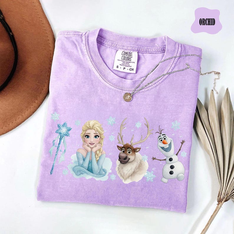 Peut inclure: T-shirt lilas avec Elsa, Olaf et Sven de La Reine des Neiges. Le t-shirt a un col rond et des manches courtes. Un collier argent&eacute; avec un pendentif est drap&eacute; sur le t-shirt. Le mot "ORCHID" est dans une bulle violette.