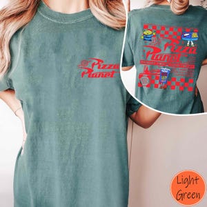 Puede incluir: Camiseta verde claro con el logo rojo "Pizza Planet" en la parte delantera. La parte trasera presenta un dise&ntilde;o a cuadros rojos y blancos con personajes de dibujos animados y texto. La camiseta es de material suave.