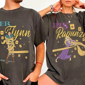 May include: Two charcoal gray t-shirts featuring character graphics. The left shirt displays Flynn Rider wrapped in golden hair with the text "HER Flynn". The right shirt shows Rapunzel with long golden hair and the text "HIS Rapunzel", both surrounded by floating lanterns.