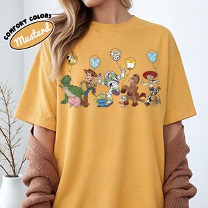 K&ouml;nnte beinhalten: Senffarbenes T-Shirt mit einer Grafik von Toy Story-Charakteren, die Luftballons halten. Zu den Charakteren geh&ouml;ren Woody, Buzz Lightyear und Rex. Das Shirt tr&auml;gt die Worte "COMFORT COLORS" und "Mustard" in einem kreisf&ouml;rmigen Design.