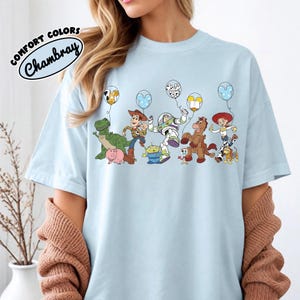 K&ouml;nnte beinhalten: Hellblaues T-Shirt mit einer bunten Grafik von Zeichentrickfiguren, die ballonf&ouml;rmige Ballons halten. Der Text "COMFORT COLORS Chambray" ist oben links aufgedruckt. Das Shirt hat einen Rundhalsausschnitt.