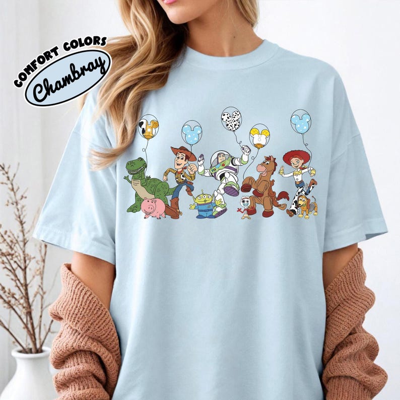 K&ouml;nnte beinhalten: Hellblaues T-Shirt mit einer bunten Grafik von Zeichentrickfiguren, die ballonf&ouml;rmige Ballons halten. Der Text "COMFORT COLORS Chambray" ist oben links aufgedruckt. Das Shirt hat einen Rundhalsausschnitt.