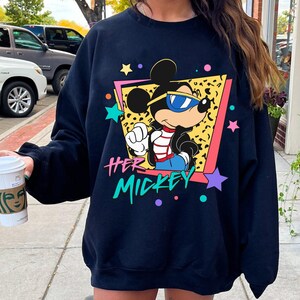 K&ouml;nnte beinhalten: Ein marineblaues Sweatshirt mit einer Grafik von Mickey Mouse mit Sonnenbrille, vor einem Leopardenmuster-Hintergrund in einem rosa und gelben Rahmen. Der Text "HER MICKEY" befindet sich darunter, mit bunten Sternen und Punkten.