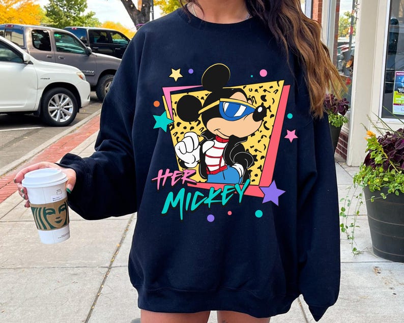 K&ouml;nnte beinhalten: Ein marineblaues Sweatshirt mit einer Grafik von Mickey Mouse mit Sonnenbrille, vor einem Leopardenmuster-Hintergrund in einem rosa und gelben Rahmen. Der Text "HER MICKEY" befindet sich darunter, mit bunten Sternen und Punkten.