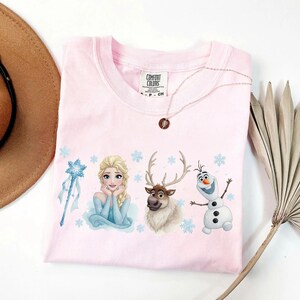 Peut inclure: Un t-shirt rose clair avec des illustrations d'Elsa, Olaf et Sven du film La Reine des Neiges, ainsi qu'un flocon de neige et une baguette. Le t-shirt a un col rond et des manches courtes. Un collier avec un pendentif est pos&eacute; sur le t-shirt.