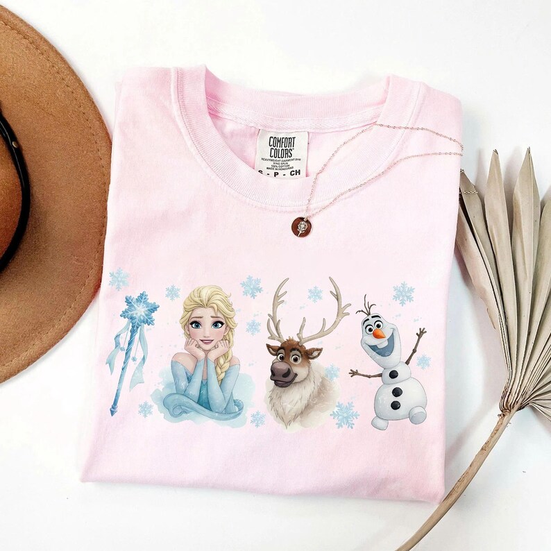 Peut inclure: Un t-shirt rose clair avec des illustrations d'Elsa, Olaf et Sven du film La Reine des Neiges, ainsi qu'un flocon de neige et une baguette. Le t-shirt a un col rond et des manches courtes. Un collier avec un pendentif est pos&eacute; sur le t-shirt.