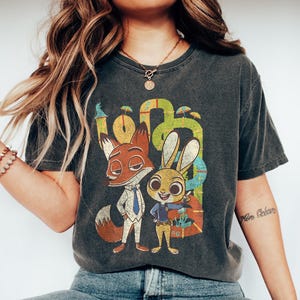 May include: Dark gray t-shirt featuring a cartoon fox and rabbit with the word "Zootopia" in colorful letters. The fox wears a suit and tie, and the rabbit is in a police uniform. The shirt has a vintage, faded look.