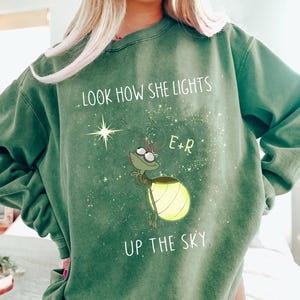 May include: Green sweatshirt with the text "LOOK HOW SHE LIGHTS UP THE SKY" and a cartoon firefly illustration. The firefly has a glowing yellow abdomen and is surrounded by stars. The sweatshirt also has the initials "E+R".