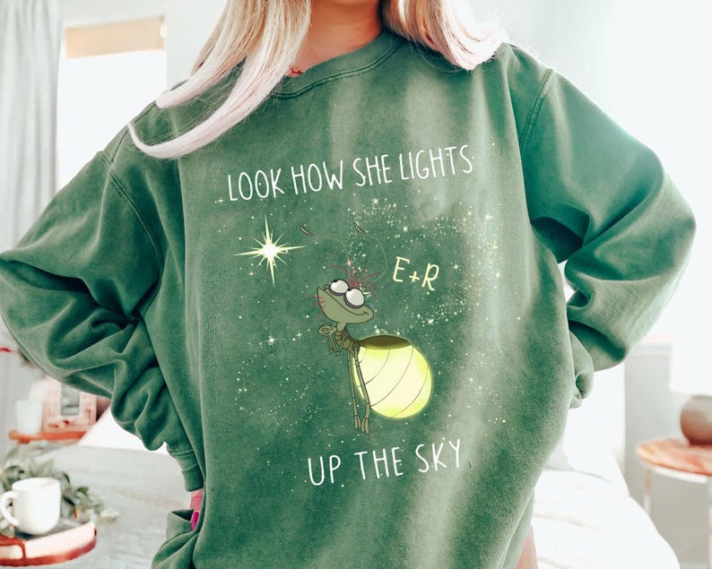 May include: Green sweatshirt with the text "LOOK HOW SHE LIGHTS UP THE SKY" and a cartoon firefly illustration. The firefly has a glowing yellow abdomen and is surrounded by stars. The sweatshirt also has the initials "E+R".