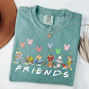 May include: A teal Comfort Colors t-shirt featuring a cartoon graphic of animal characters holding Mickey Mouse-shaped balloons. The word "FRIENDS" is printed in white block letters across the bottom. A silver necklace with a pendant is draped across the shirt.