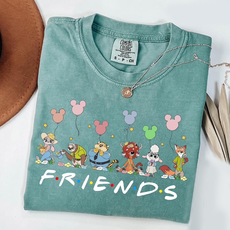 May include: A teal Comfort Colors t-shirt featuring a cartoon graphic of animal characters holding Mickey Mouse-shaped balloons. The word "FRIENDS" is printed in white block letters across the bottom. A silver necklace with a pendant is draped across the shirt.