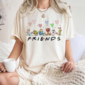 May include: Cream-colored t-shirt featuring the word "FRIENDS" in black block letters. Above the word, there is a cartoon illustration of several animal characters holding colorful balloons. The shirt is a casual, unisex style.