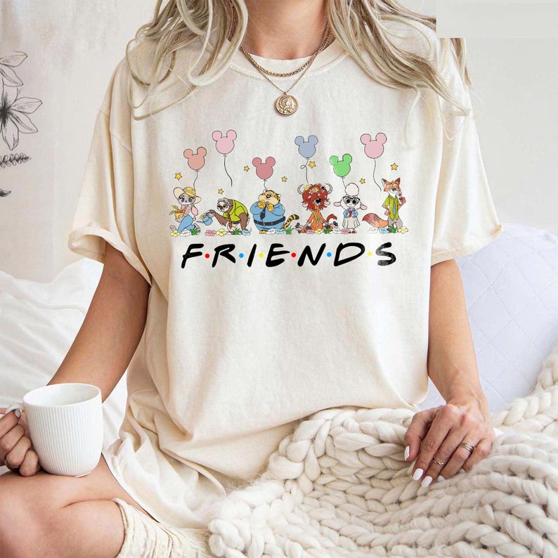 May include: Cream-colored t-shirt featuring the word "FRIENDS" in black block letters. Above the word, there is a cartoon illustration of several animal characters holding colorful balloons. The shirt is a casual, unisex style.