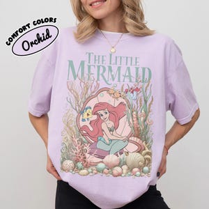 May include: Orchid-colored t-shirt featuring a graphic of Ariel from The Little Mermaid. The design includes the text "The Little Mermaid" in green, with Ariel inside a seashell, surrounded by sea creatures and coral.