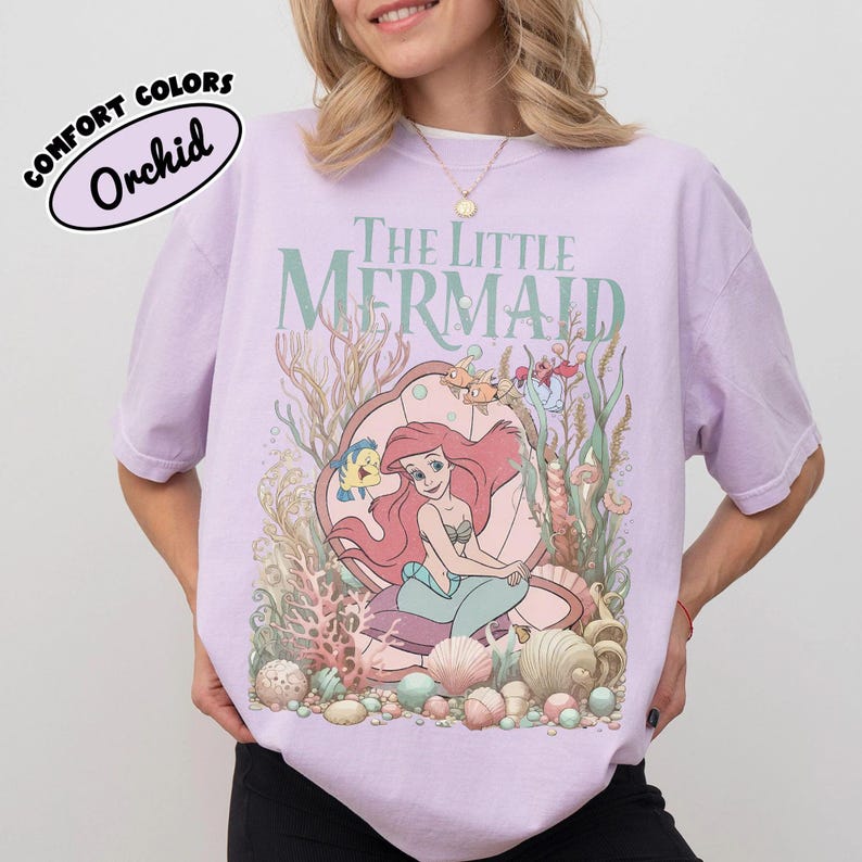 May include: Orchid-colored t-shirt featuring a graphic of Ariel from The Little Mermaid. The design includes the text "The Little Mermaid" in green, with Ariel inside a seashell, surrounded by sea creatures and coral.