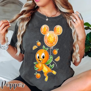 Puede incluir: Camiseta gris oscuro con un personaje de dibujos animados de naranja sosteniendo una naranja y un globo con forma de Mickey Mouse. El diseño incluye naranjas y hojas. La palabra "Pepper" está impresa en la parte inferior izquierda.