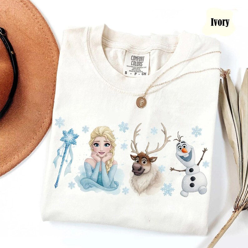 Peut inclure: T-shirt ivoire avec Elsa, un renne et Olaf de La Reine des Neiges. Le t-shirt a un col rond et des manches courtes. Un collier en or avec un pendentif est drap&eacute; sur le t-shirt. Le t-shirt est &eacute;tiquet&eacute; "Comfort Colors".
