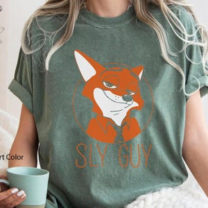 Puede incluir: Camiseta de color verde musgo con un zorro de dibujos animados naranja y el texto "SLY GUY". El zorro est&aacute; dentro de un c&iacute;rculo. La camiseta es de manga corta y cuello redondo.