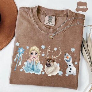 Peut inclure: T-shirt couleur expresso avec un motif d'Elsa, d'un renne et d'Olaf du film La Reine des Neiges. Le t-shirt a un col rond et des manches courtes. Le texte "Comfort Colors" est visible sur l'&eacute;tiquette.