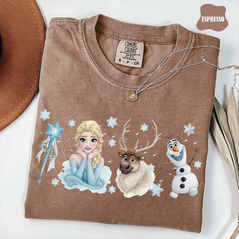 Peut inclure: T-shirt couleur expresso avec un motif d'Elsa, d'un renne et d'Olaf du film La Reine des Neiges. Le t-shirt a un col rond et des manches courtes. Le texte "Comfort Colors" est visible sur l'&eacute;tiquette.