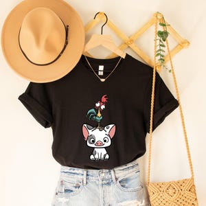 Puede incluir: Camiseta negra con una ilustraci&oacute;n de dibujos animados de un gallo de pie sobre un cerdo. La camiseta se combina con pantalones cortos de mezclilla. Tambi&eacute;n se ven un sombrero beige, un collar dorado y una bolsa tejida.