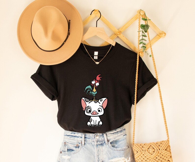 Puede incluir: Camiseta negra con una ilustraci&oacute;n de dibujos animados de un gallo de pie sobre un cerdo. La camiseta se combina con pantalones cortos de mezclilla. Tambi&eacute;n se ven un sombrero beige, un collar dorado y una bolsa tejida.