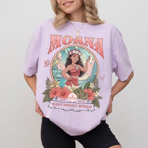 May include: A light purple t-shirt featuring the word "MOANA" in a retro font. The shirt has a graphic of Moana with flowers and the text "WALT DISNEY WORLD" below. The shirt is made of a soft material.