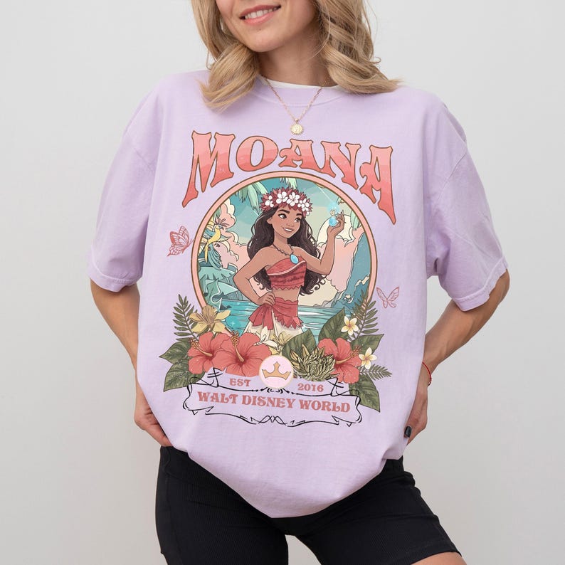 May include: A light purple t-shirt featuring the word "MOANA" in a retro font. The shirt has a graphic of Moana with flowers and the text "WALT DISNEY WORLD" below. The shirt is made of a soft material.