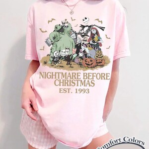 Puede incluir: Camiseta rosa claro con un dise&ntilde;o de "Pesadilla antes de Navidad". El dise&ntilde;o incluye personajes como Jack Skellington, Sally y Oogie Boogie, con murci&eacute;lagos y calabazas. El texto "NIGHTMARE BEFORE CHRISTMAS EST. 1993" est&aacute; impreso debajo.