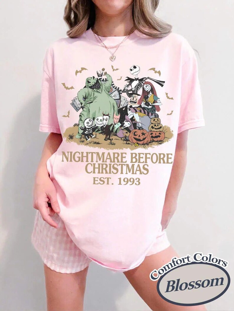 Puede incluir: Camiseta rosa claro con un dise&ntilde;o de "Pesadilla antes de Navidad". El dise&ntilde;o incluye personajes como Jack Skellington, Sally y Oogie Boogie, con murci&eacute;lagos y calabazas. El texto "NIGHTMARE BEFORE CHRISTMAS EST. 1993" est&aacute; impreso debajo.